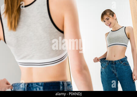 happy slim woman in oversized jeans looking at mirror isolated on white Stock Photo