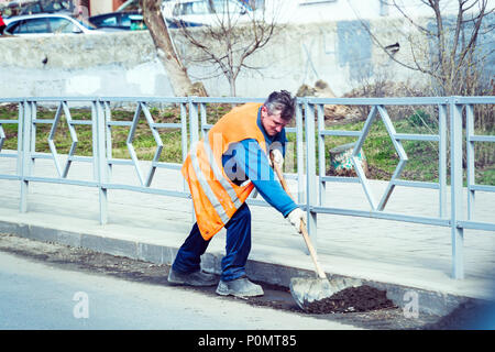 The janitor in an orange uniform cleans the garbage with a shovel on the city street Stock Photo