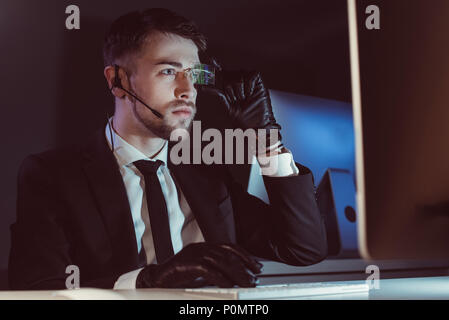 portrait of hacker with headset looking at computer screen at table in dark Stock Photo