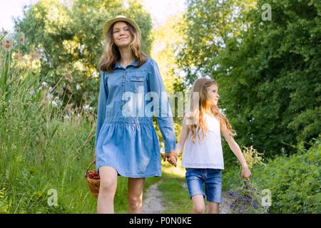 Children girls on forest road holding hands. Sunny summer day, girl holding basket with berries. Stock Photo