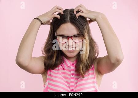 Teen girl with glasses, with long hair scratches her head and is emotional puzzled. Pink studio background. Stock Photo