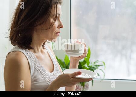 Mature woman drinks morning coffee and looks out the window at home. Photo in profile female serious Stock Photo