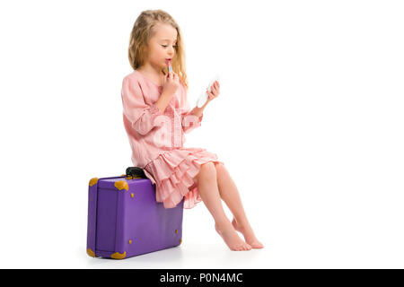 Side view of stylish child sitting on suitcase and doing makeup by lipstick isolated on white Stock Photo