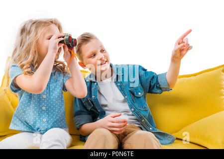 boy pointing while sister looking at binoculars, sitting on yellow sofa Stock Photo