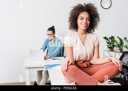 Businesswoman sitting in lotus position in office with male coworker working on laptop behind Stock Photo