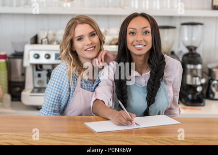 attractive multiethnic waitresses smiling and writing an order at cafe Stock Photo