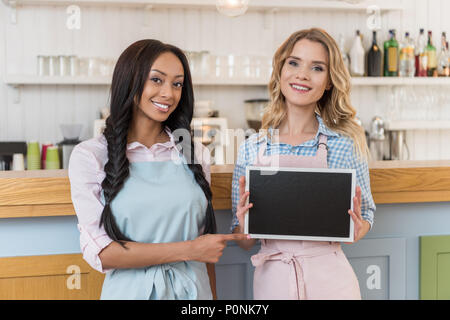 smiling beautiful multiethnic waitresses with blank board looking at camera while standing at cafe Stock Photo
