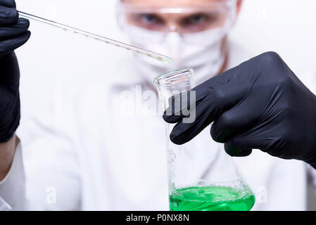 The scientist's hands hold a flask and a test tube with a bright green liquid. The experiment is conducted by the scientist in protective glasses, a m Stock Photo