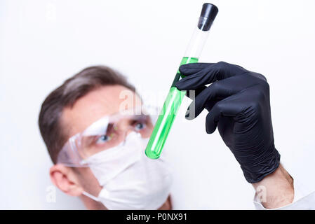 A test tube with a green liquid in the hands of a scientist wearing protective goggles and a mask on a white background. Stock Photo