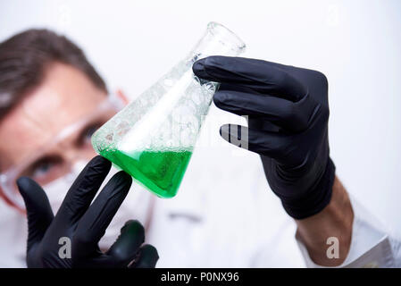 A flask with a green liquid and bubbles in the hands of a young man. The scientist in uniform in protective glasses, a mask and black gloves conducts  Stock Photo