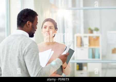 Bearded African American entrepreneur studying statistic data with young subordinate while standing at modern open plan office, lens flare Stock Photo
