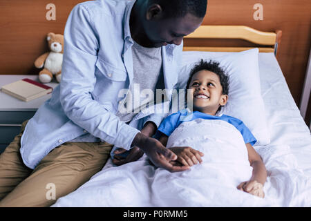 smiling african american boy looking at father while lying in bed in clinic Stock Photo