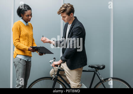 handsome young businessman signing contract in hands of young african american colleague while sitting on bike Stock Photo