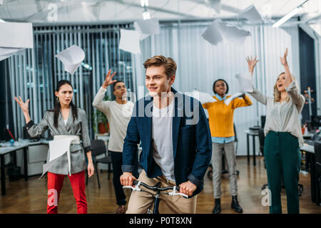 young businessman riding bike at office while partners throwing up papers on background Stock Photo