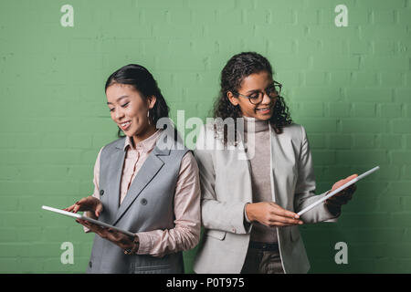 african american and asian businesswomen using digital tablets in front of green wall Stock Photo