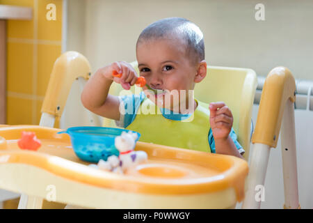 The boy 2 years eats porridge. Children's table. The concept of the child's independence. funny kid in a baby seat Stock Photo