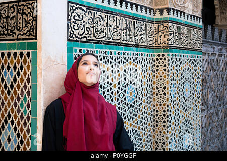 Muslim woman in traditional clothing with red hijab and black dress in front of traditional arabesque decorated wall with text from Koran Stock Photo