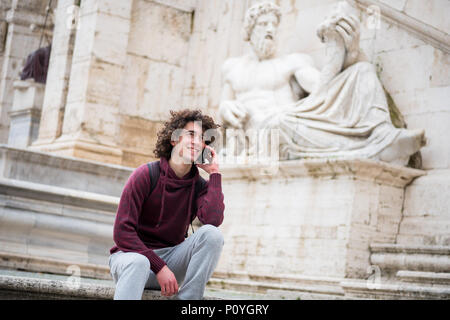 Handsome young man with curly hair in tracksuit talking on his mobile phone in front of Nile God statue in Rome Stock Photo