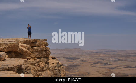 Young man standing on cliff edge and taking pictures of the desert on his phone Stock Photo