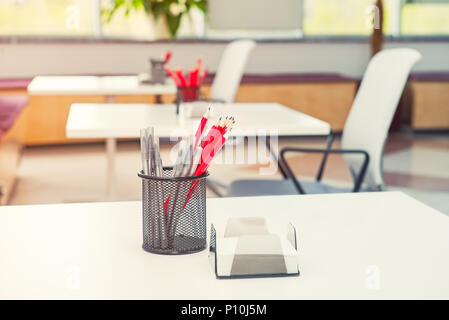 Close up desk with focused stand with stationery, pens and pencils with blurred background of modern light empty open space office. Coworking workplac Stock Photo