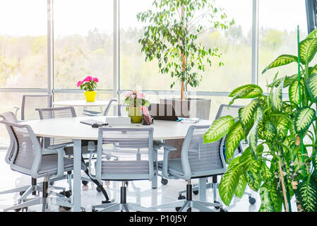 Round table as work place with laptop, notebooks in modern light open space office with big windows, desks, chairs and green plants. Coworking. Minima Stock Photo