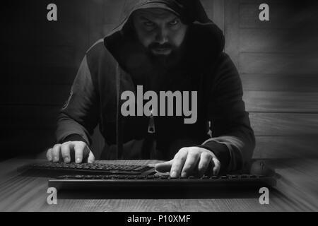 A bearded man in a hood sits and works behind a computer hacker Stock Photo
