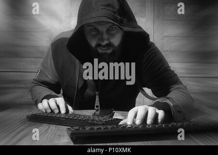 A bearded man in a hood sits and works behind a computer hacker Stock Photo