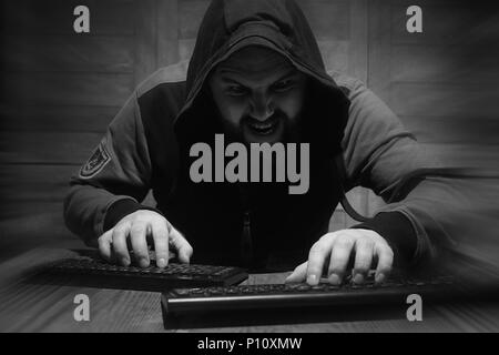A bearded man in a hood sits and works behind a computer hacker Stock Photo
