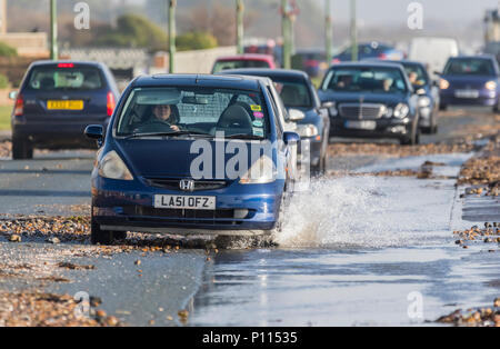 Car driving slowly through water on flooded road after heavy rain and high tide sea water and pebbles from the beach onto the road in West Sussex, UK. Stock Photo