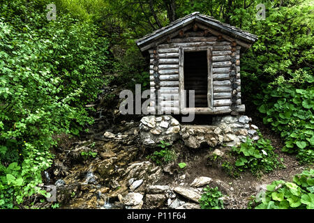 tiny wooden mountain cabin in dense lush green summer forest with a small creek beside it Stock Photo