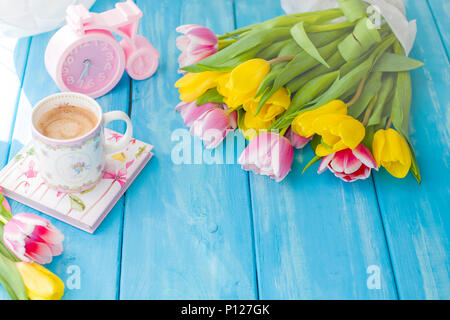 A cup of coffee on a notebook on a blue wooden background. Bright colors. Bouquet of flowers yellow and pink. The pink clock is like a bicycle. Spring morning. Text. View from above Stock Photo