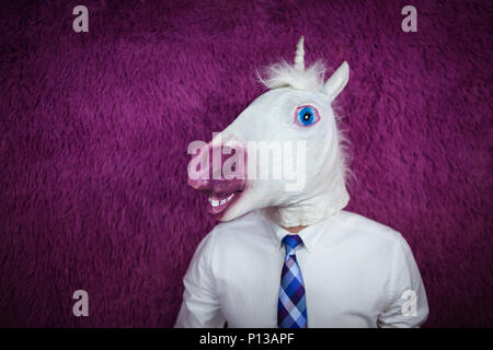Freaky young man in comical mask stands on the purple background. Portrait of unusual manager. Serious unicorn in shirt and tie is looking away Stock Photo