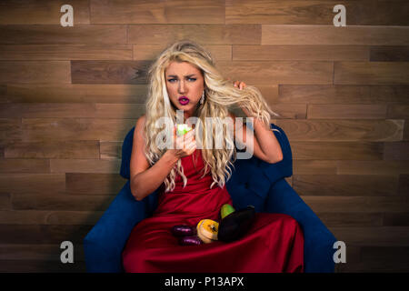 Young vegetarian woman in elegant red dress sits on armchair and holds food in hands. Unhappy girl holds raw vegetables and eats vegetables Stock Photo