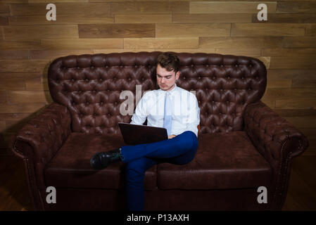 Serious man in suit is working with laptop at home office. Confident young manager sits on the leather sofa on background of wooden wall. Stock Photo