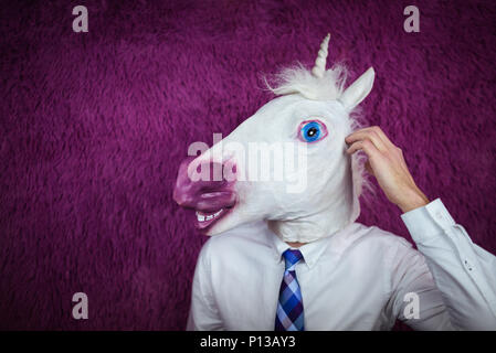Freaky young man in comical mask stands on the purple background. Portrait of unusual manager. Confused unicorn in shirt and tie is thinking Stock Photo