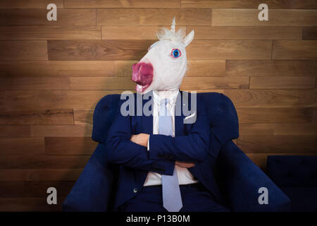 Portrait of unusual man at home office. Freaky young manager in comical mask on background of wooden wall. Unicorn in suit sits on armchair like boss Stock Photo