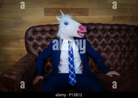 Portrait of unusual man at home office. Freaky young manager in comical mask on background of wooden wall. Unicorn in suit sits on sofa like a boss Stock Photo