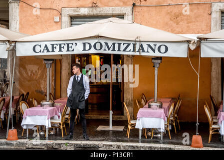 Italian waiter stands outside Caffe Domiziano restaurant on Piazza Navona waiting for customers on a rainy day with empty tables for outdoor dining. Stock Photo