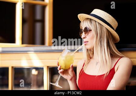 Young pretty sympathetic blonde sitting in a cafe near an open window with a cocktail in her arms and in a straw hat. Stock Photo