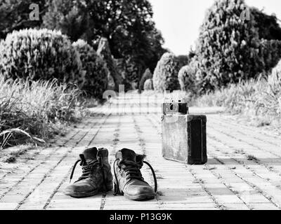 Leather boots in a foreground, in the middle of a brick road. A film camera on an antique suitcase, in a blurry background. Black-and-white photo. Stock Photo