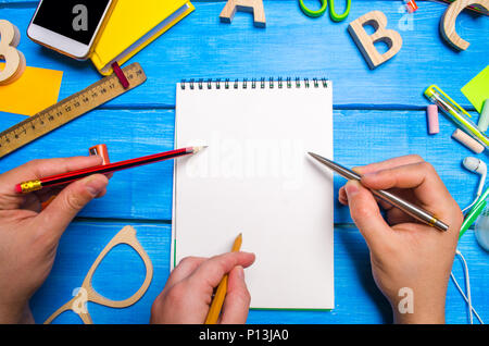 Three hands with pencils and pens point to a notebook with white pages on the desktop blue wood background. Creative disorder. People develop new idea Stock Photo