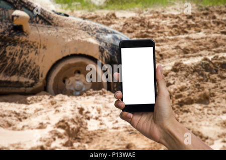 Man hand holding smartphone with white screen display use for mock up. Car tire stuck in the mud background. Car wheel in the mud clay can not run out Stock Photo