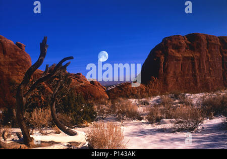 Arches National Park: view of distant horizon through rock formations with full moon over snow covered desert in late light, near Moab, Utah, USA Stock Photo