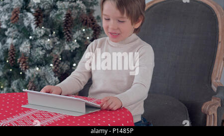 Cute little boy using tablet while sitting on a big armchair at home over Chirstmas tree background Stock Photo