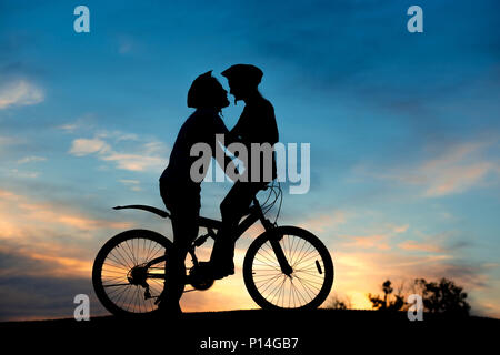 Romantic couple of cyclists is kissing at sunset. Young man and woman is kissing on bike at evening sky background. Enjoying of each other. Stock Photo