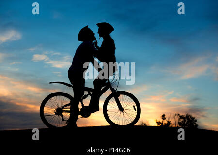 Couple kissing on the top of hill at sunset. Silhouette of bicyclists is kissing on summer nature background with beautiful clouds in the sky. Romanti Stock Photo