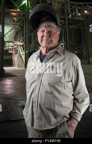 Strong senior man with a welding helmet on standing in front of an industrial building at night with lights showing in the building Stock Photo