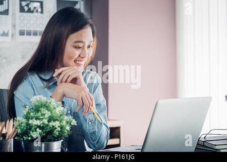 Asian businesswoman thinking with pencil and looking down to laptop computer screen for planning work with smile face in office,Office lifestyle conce Stock Photo