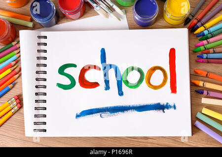 The word School painted on a white art book with various paints, crayons and pencils on a school desk.