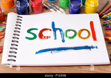 The word School painted on a white art book with various paints, crayons and pencils on a school desk.